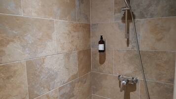 Un baño con azulejos color crema, un cabezal de ducha manual, un grifo empotrado en la pared y una botella de jabón líquido.