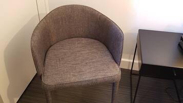 Una silla con tela texturizada y patas metálicas, ubicada junto a una pared blanca y un pequeño escritorio con estructura negra.