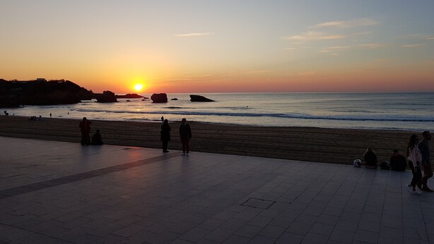 A beach with people walking and sitting, a sunset, and rocky formations in the water. - Hôtel Au Bon Coin (Biarritz)
