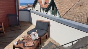 A rooftop terrace with a wooden deck, a cushioned chair, a small table with food and drinks, and a view of the cityscape. - Hôtel Au Bon Coin (Biarritz)