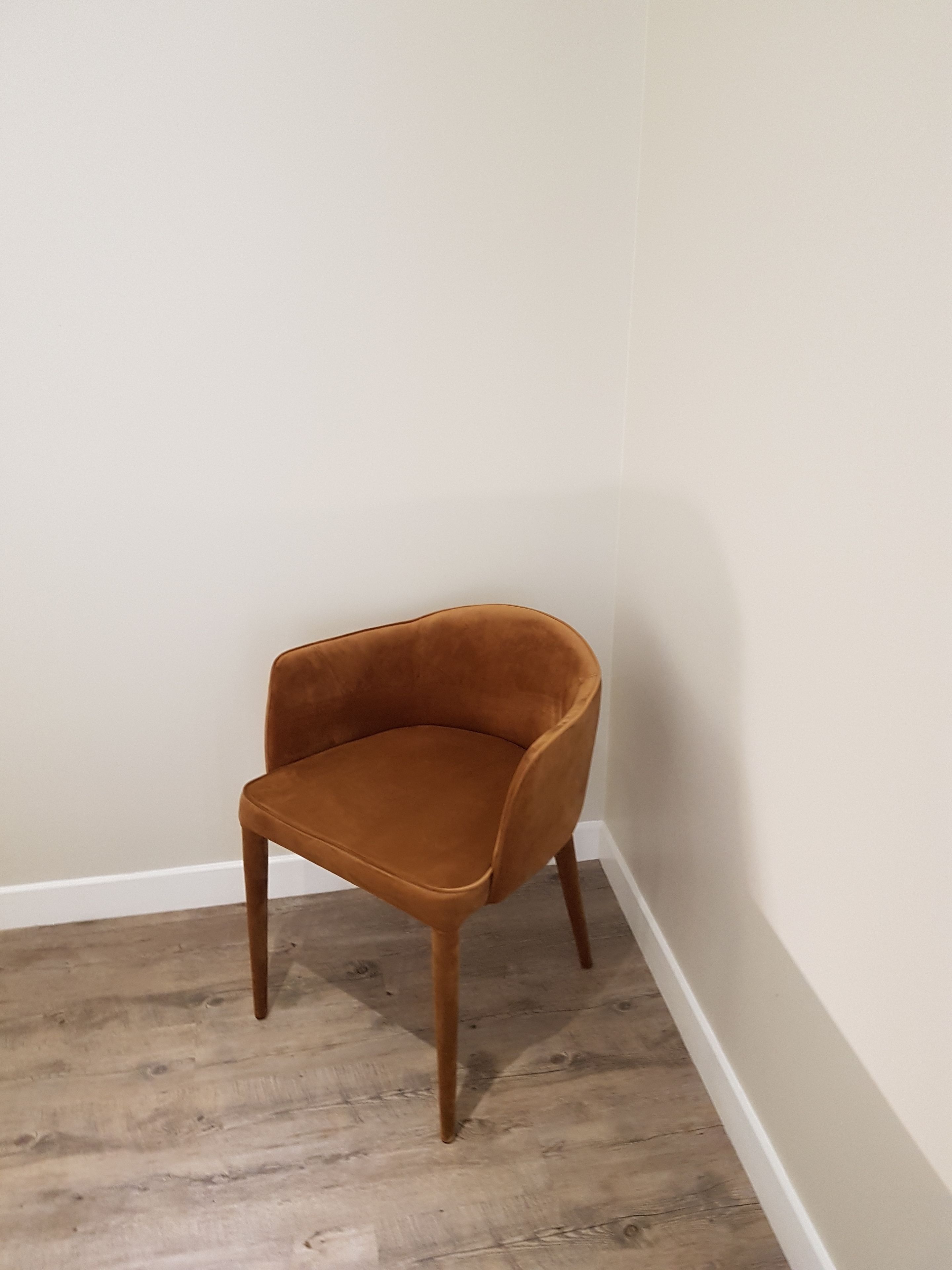 Una silla marrón individual con patas de madera en un rincón de una habitación con suelo de madera y paredes blancas.