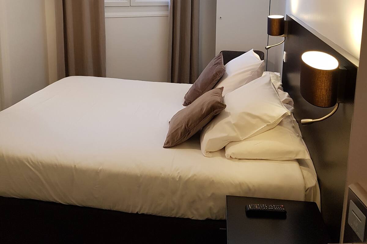 A hotel room with a bed, a nightstand, a lamp, a window with curtains, and a door.