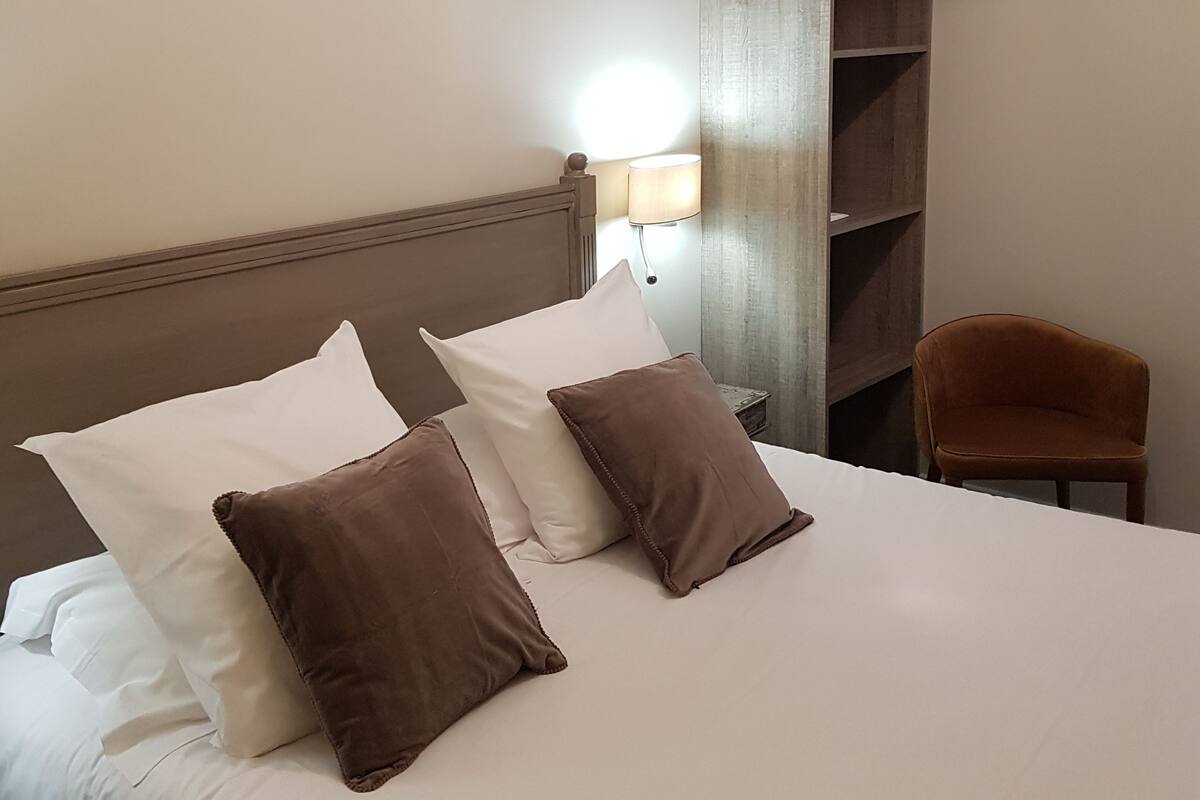 A hotel room with a bed, pillows, a nightstand, a lamp, a chair, and a shelf.