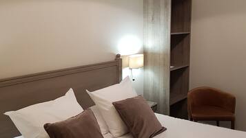 Una habitación de hotel con cama, almohadas, mesita de noche, lámpara, silla y estante.