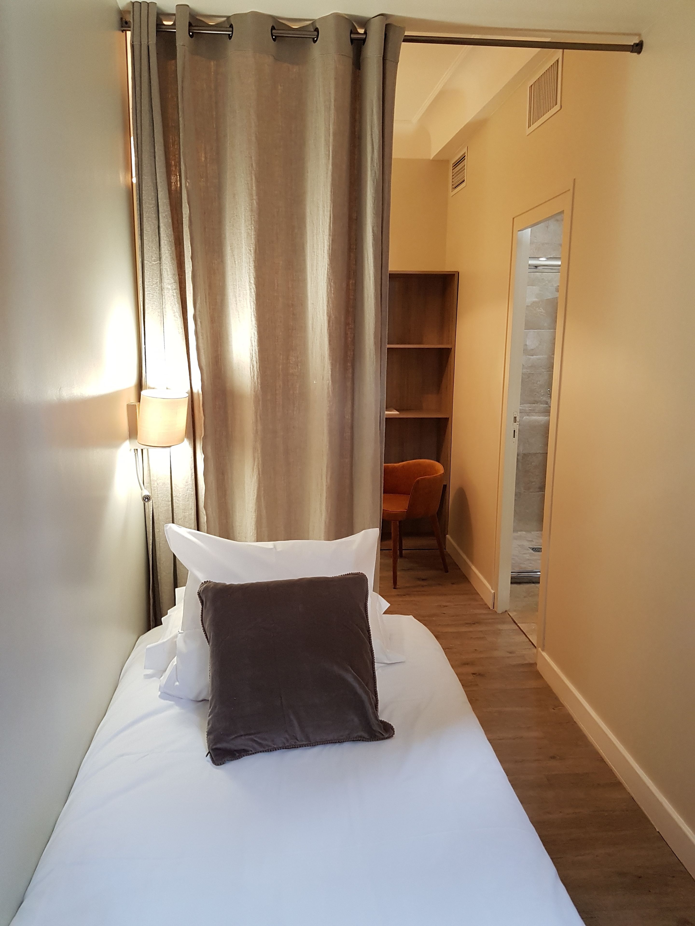 A narrow room with a bed, a chair, and a wooden shelf unit.