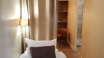 A narrow room with a bed, a chair, and a wooden shelf unit.