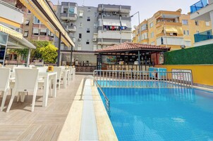 A rooftop pool area with white chairs and tables, a bar, and a view of surrounding buildings.