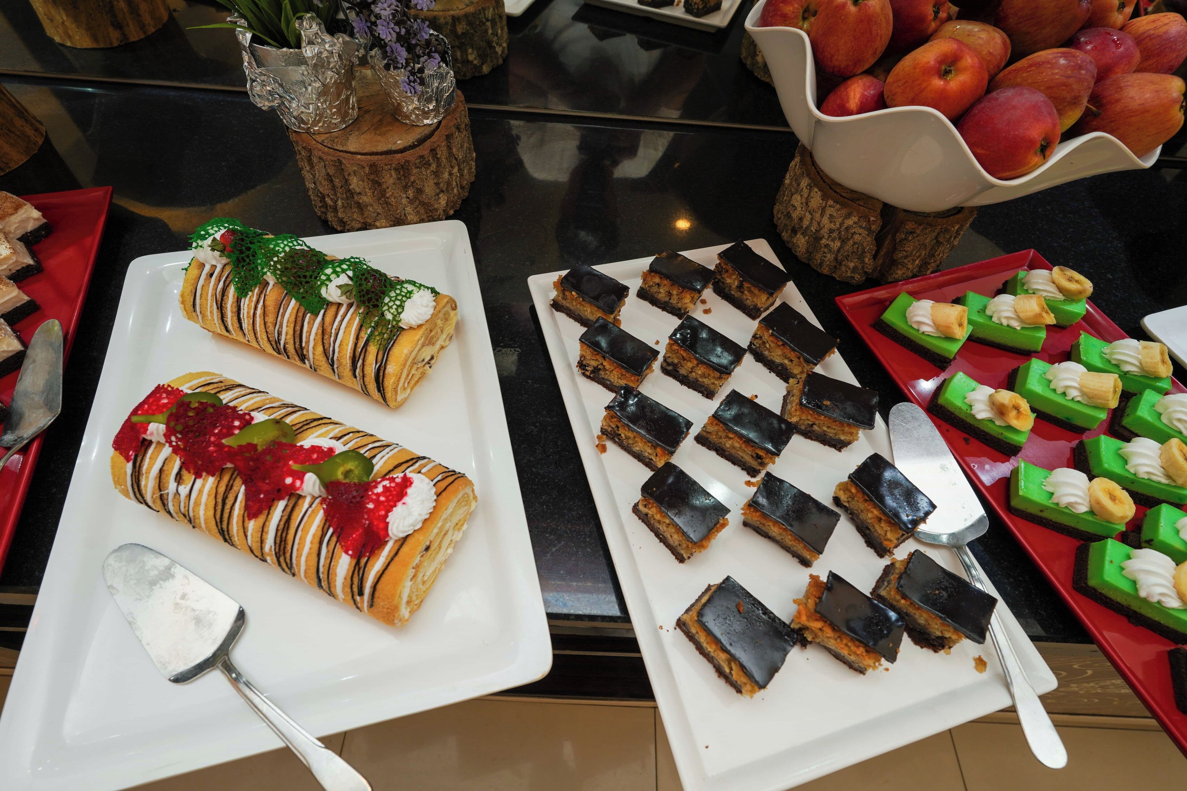 A buffet table with various desserts, including a rolled pastry with red fruit and white cream, chocolate-topped squares, and green desserts with banana slices.