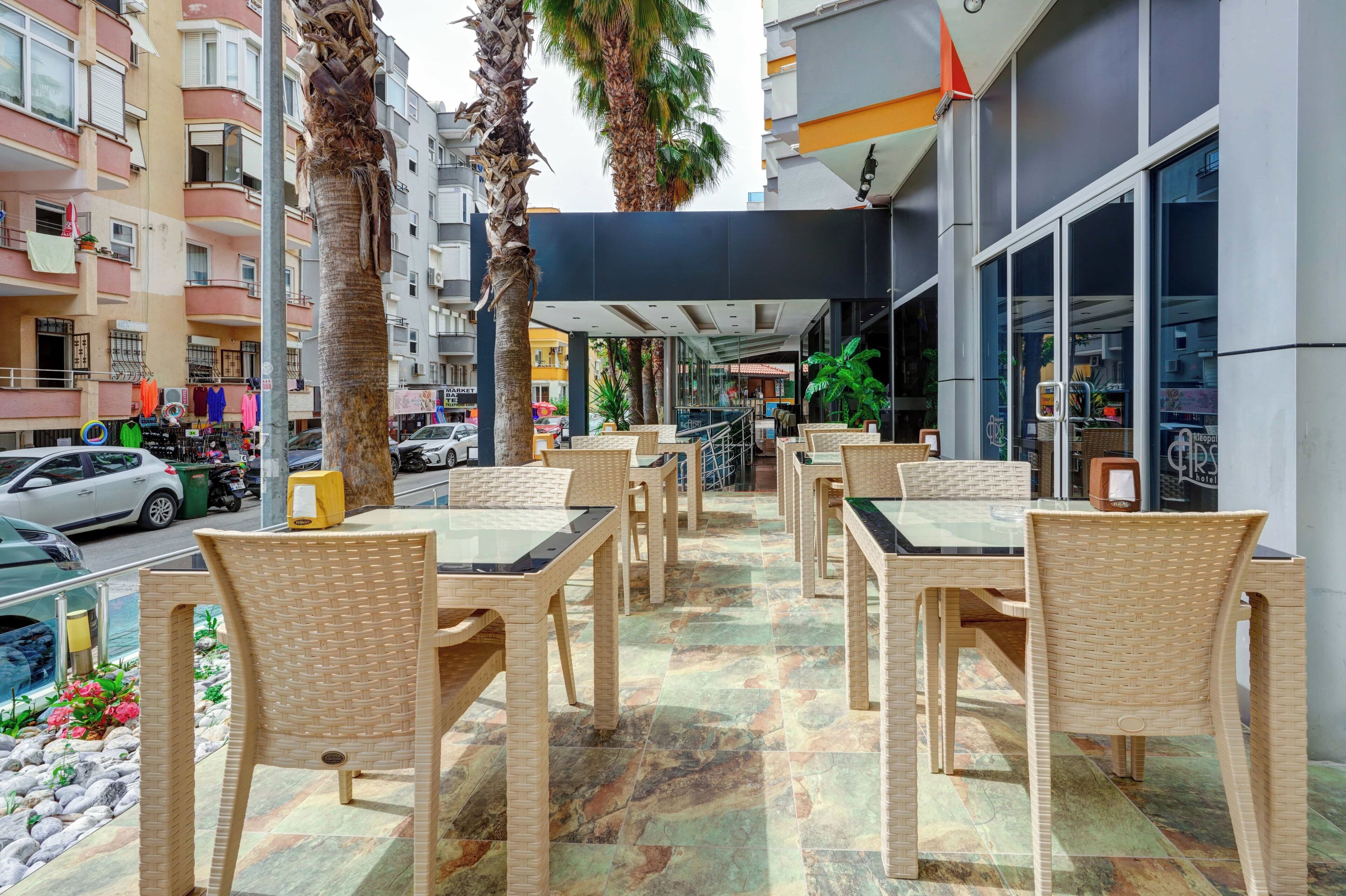 An outdoor seating area with wicker chairs and tables, surrounded by palm trees and a view of a street with parked cars and buildings.
