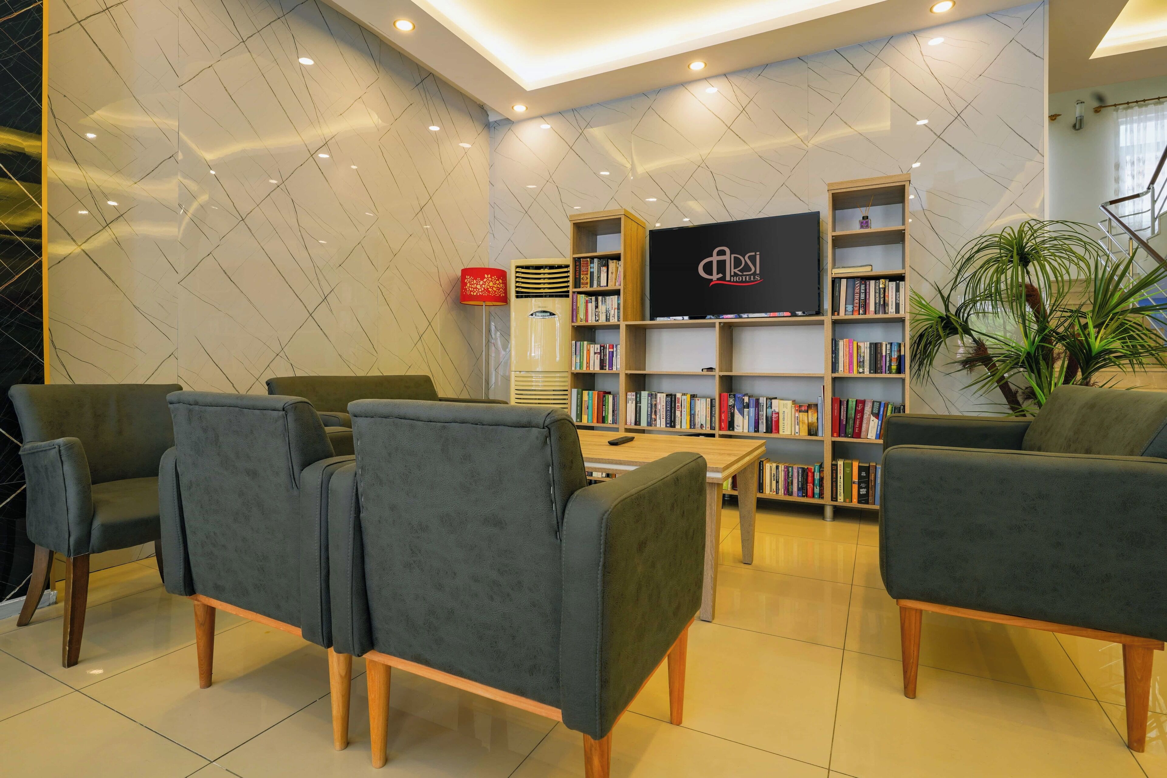 A modern hotel lobby with grey upholstered chairs, a flat-screen TV, and a bookshelf filled with books.
