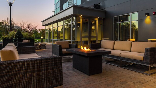 An outdoor seating area with wicker furniture and a central fire feature, adjacent to a modern building with large windows.