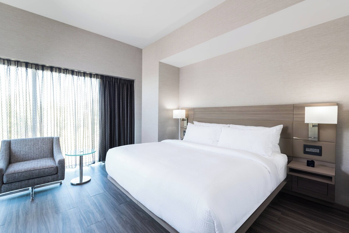 A hotel room with a large bed, a chair, a small table, and a nightstand with a lamp.