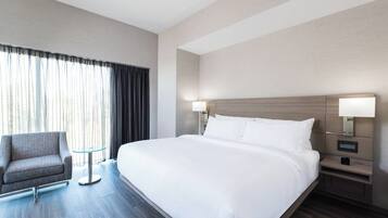 A hotel room with a large bed, a chair, a small table, and a nightstand with a lamp.