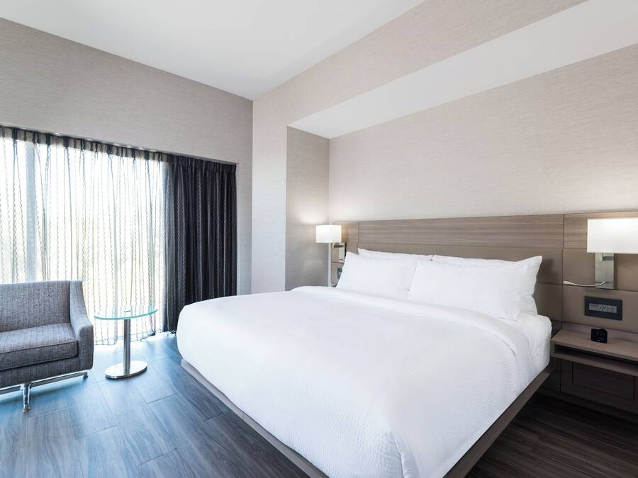 A hotel room with a large bed, a chair, a small table, and a nightstand with a lamp.