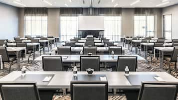 Un salón de conferencias con varias mesas y sillas, una pantalla grande al frente y piso alfombrado.