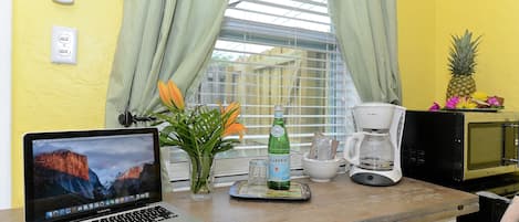 A laptop on a wooden table with a window, curtains, a pineapple, a bottle, and a coffee maker.