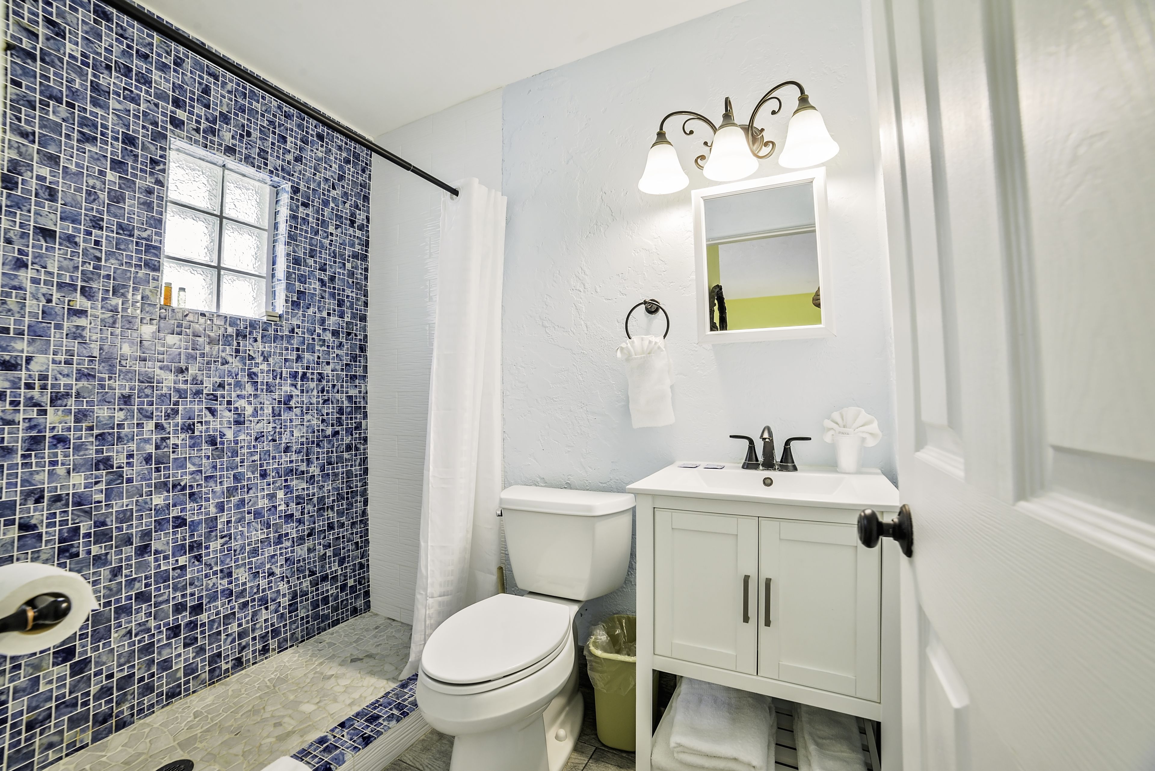 Une salle de bain avec des toilettes blanches, un lavabo avec un miroir et un espace douche aux carreaux de mosaïque bleus.