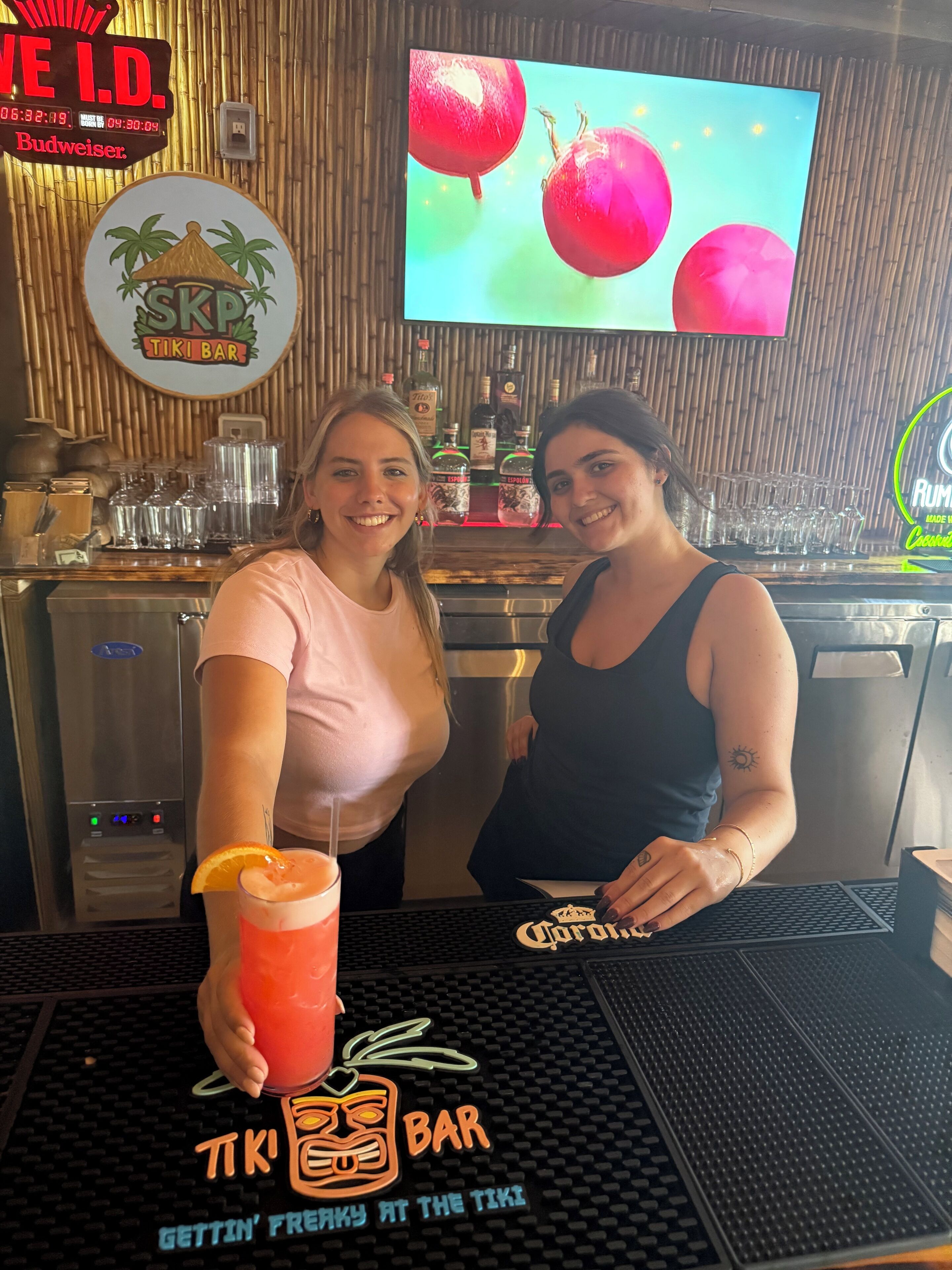 Two people at a bar with a tropical theme, including a sign for 'SKP TIKI BAR' and a television displaying fruit.