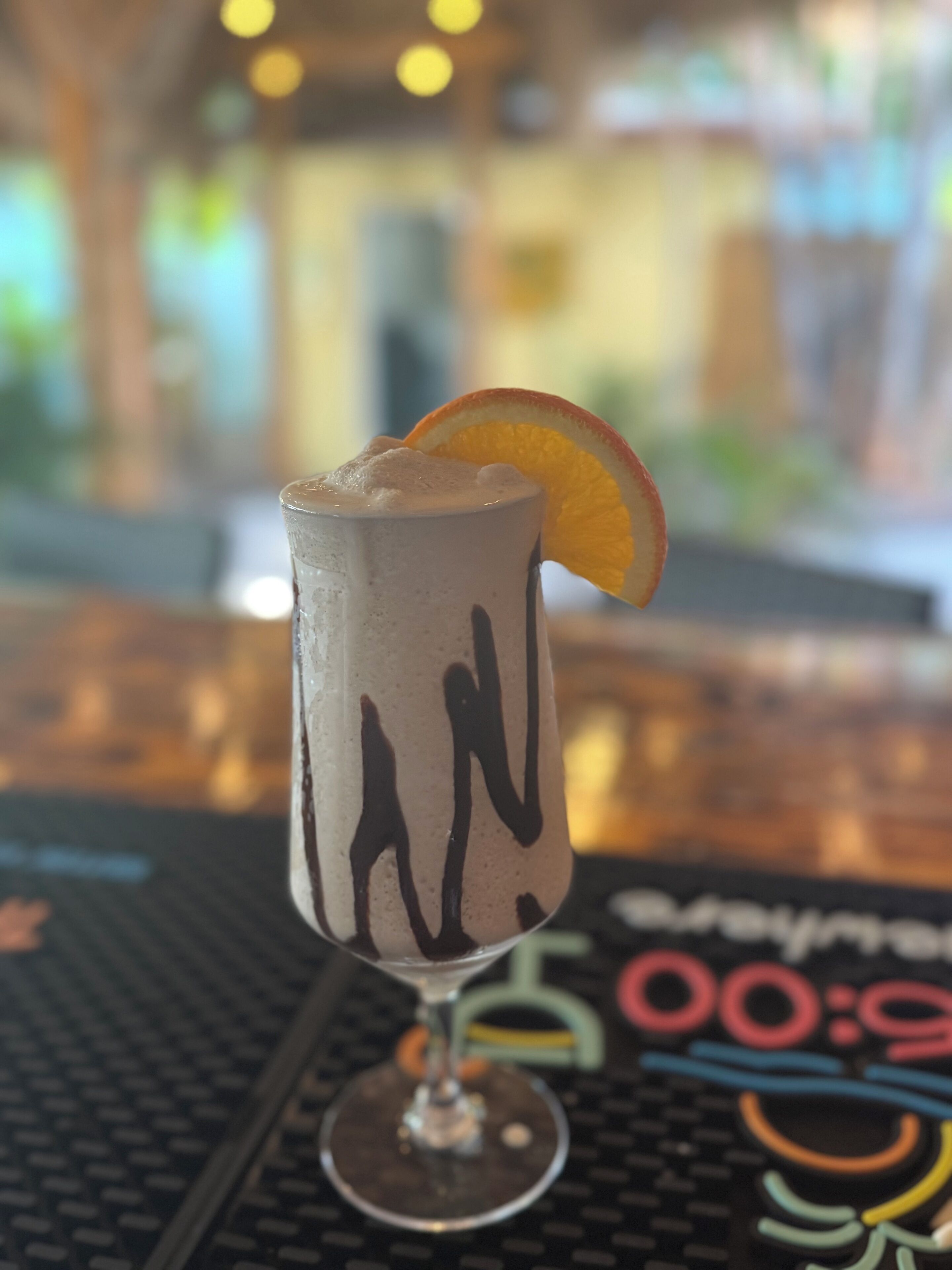 A cocktail glass with a frothy drink, a slice of orange, and a black drizzle on the rim, placed on a bar mat with a blurred background.