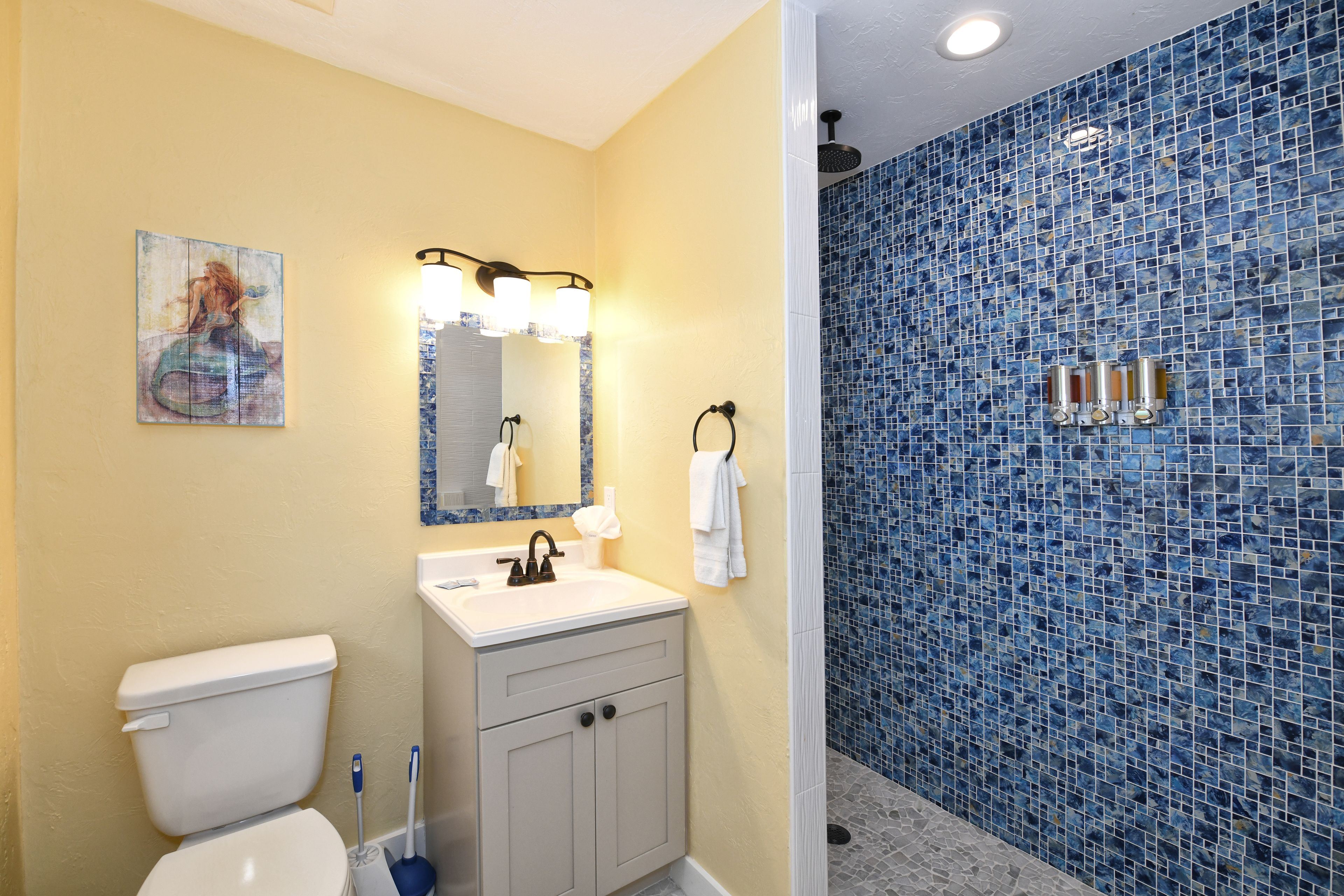 A bathroom with a toilet, sink, and mirror. There is a shower with blue mosaic tiles.