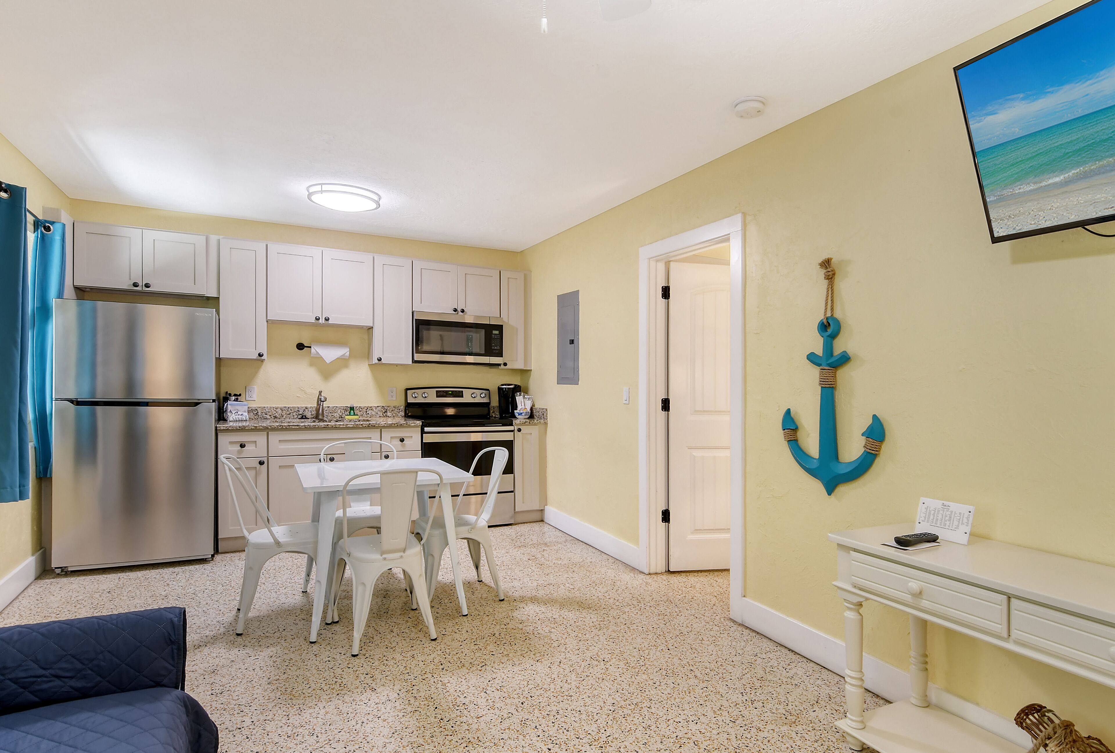A compact kitchen with white cabinets, a stainless steel refrigerator, and a microwave. There is a dining area with a table and chairs, and a TV mounted on the wall.