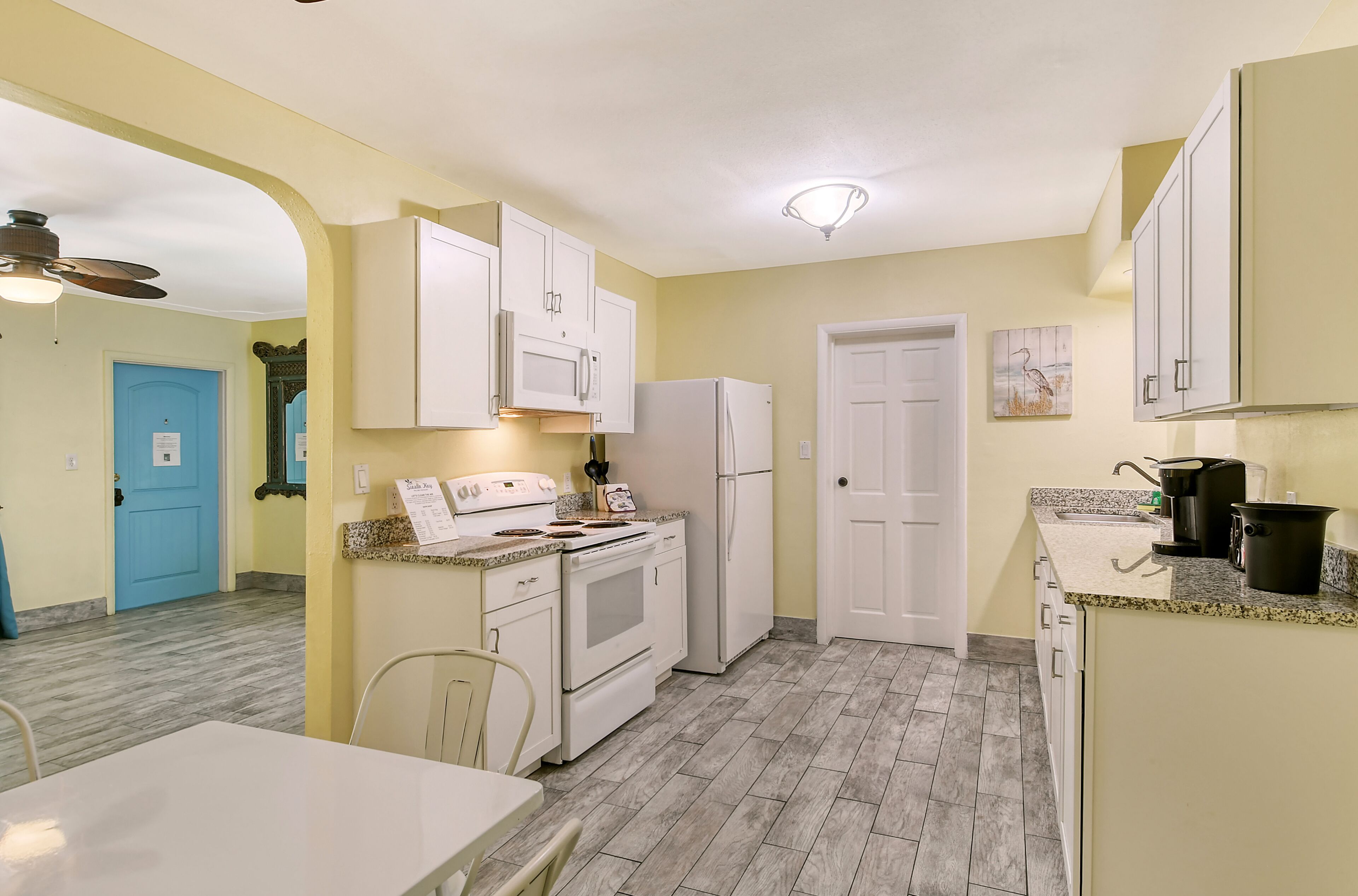 A compact kitchen with white cabinets, a stove, and a refrigerator. There is a dining table with chairs and a blue door in the background.