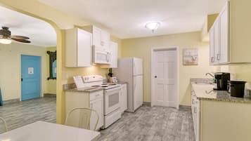 Cocina pequeña con armarios blancos, estufa y refrigerador. Se ve una mesa de comedor con sillas y una puerta azul al fondo.