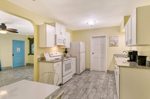 Cocina pequeña con armarios blancos, estufa y refrigerador. Se ve una mesa de comedor con sillas y una puerta azul al fondo.