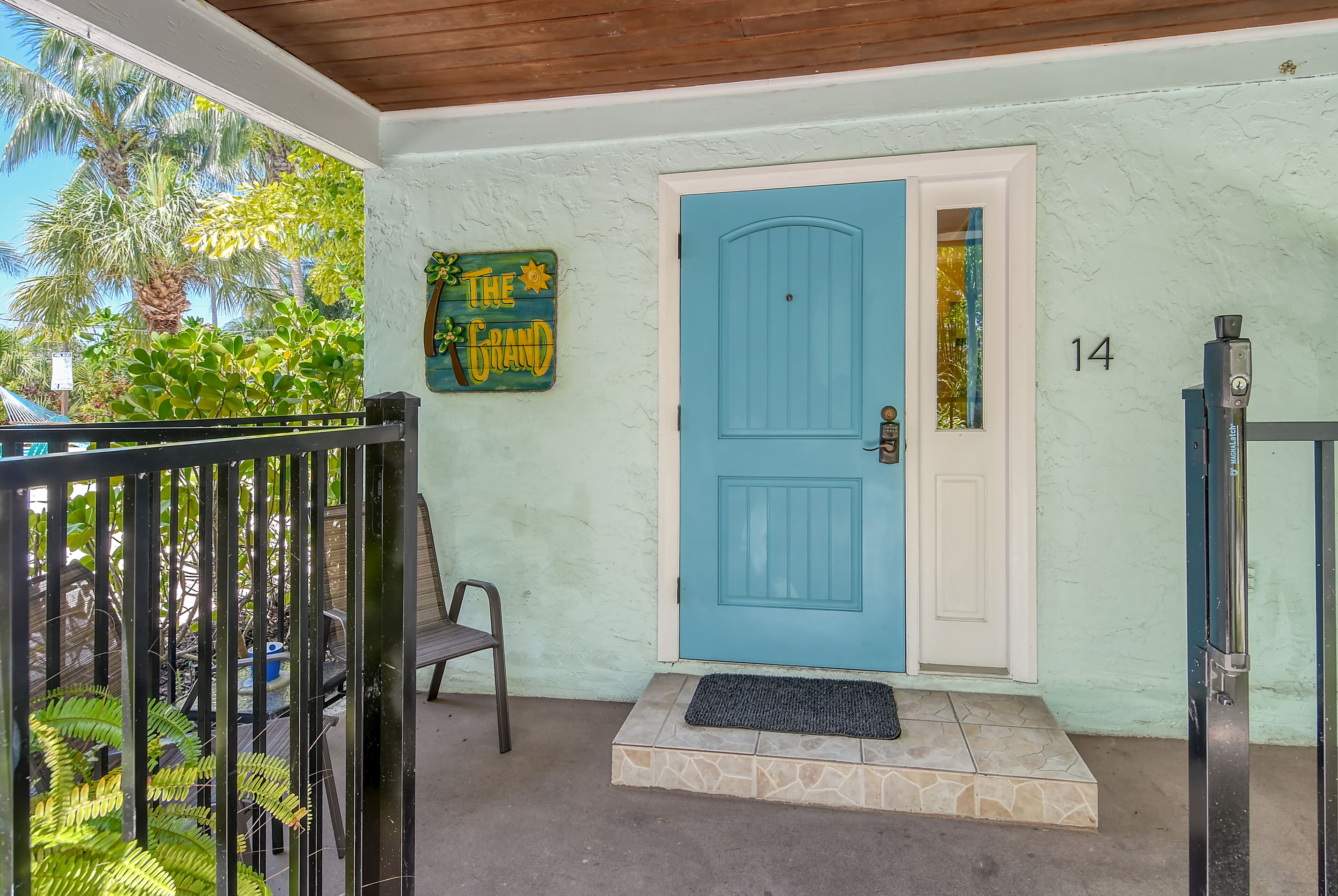 A blue door with a white panel, a number '14' on the wall, a black gate, a chair, and a sign that reads 'The Grand'.