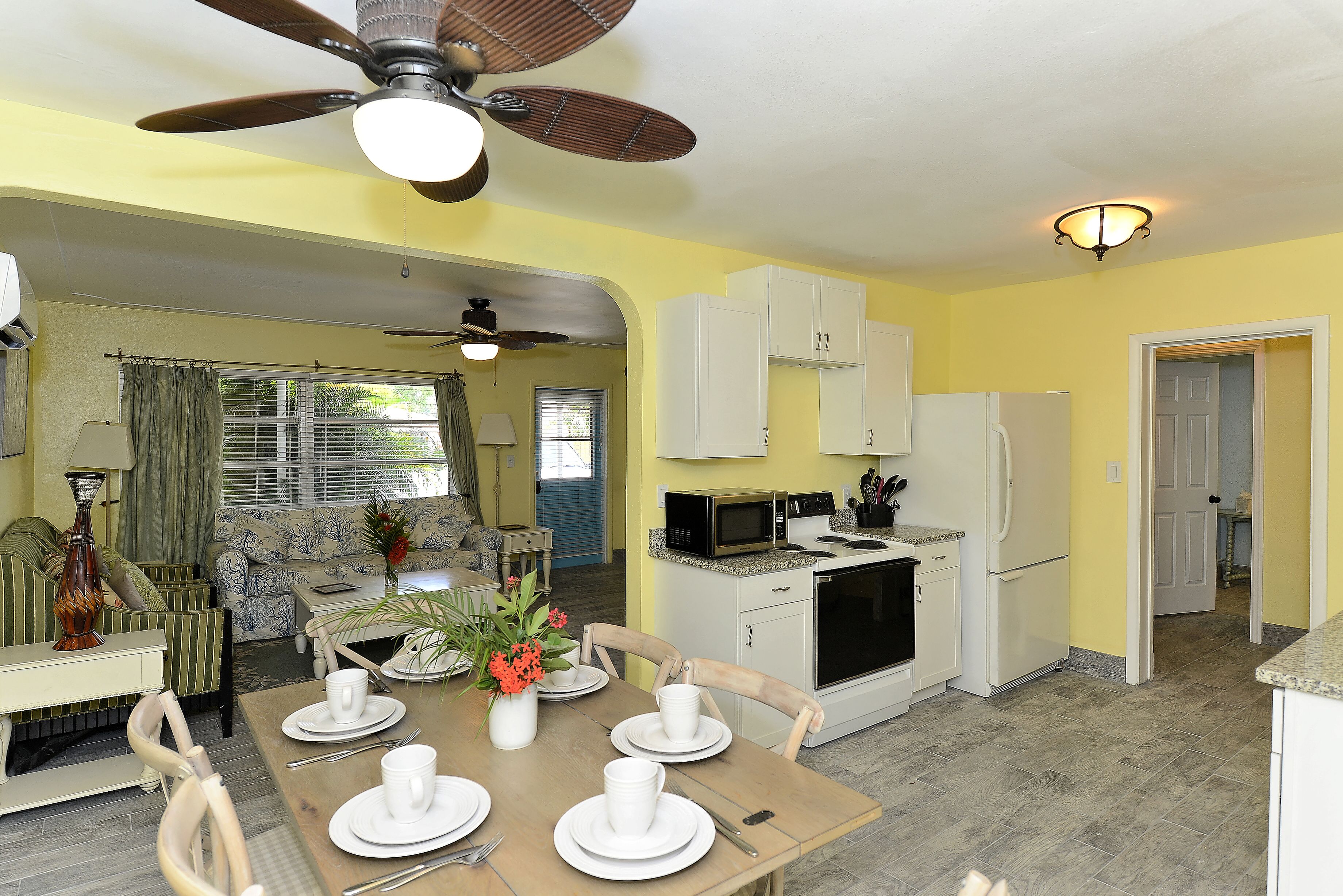 A dining area with a wooden table set for four, white plates, and a vase with flowers. There is a kitchen with white cabinets and appliances, and a ceiling fan above the dining table.