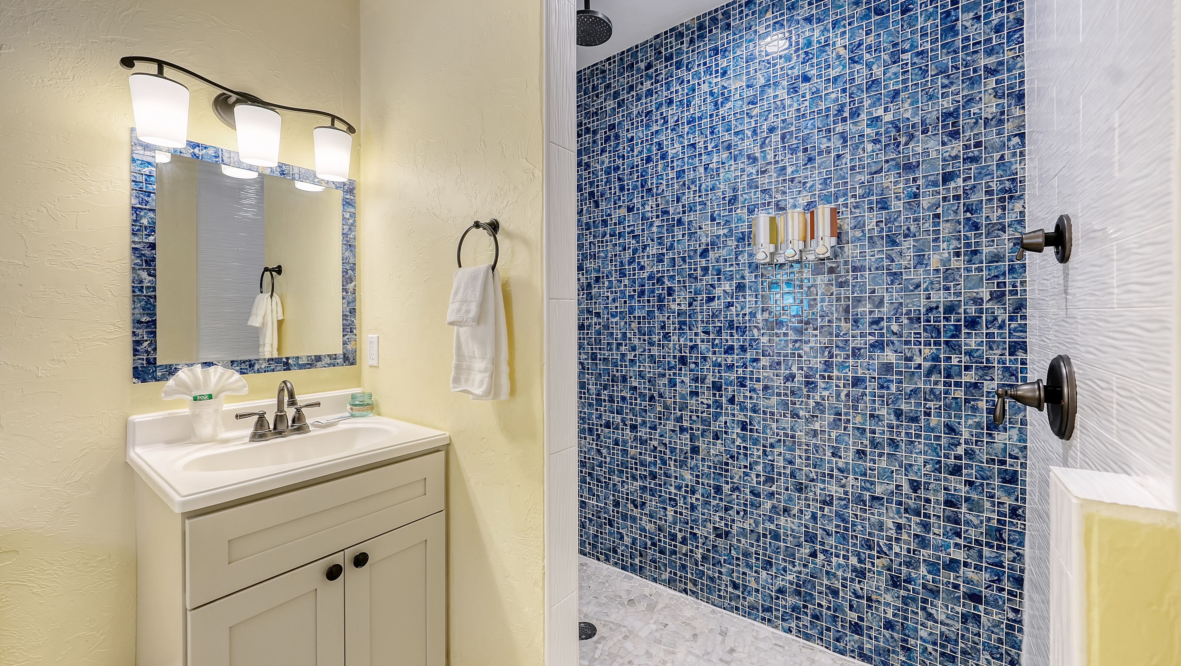 Un baño con lavamanos, espejo y tocador blancos, y una ducha con mosaicos azules.