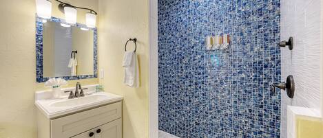 Un baño con lavamanos, espejo y tocador blancos, y una ducha con mosaicos azules.