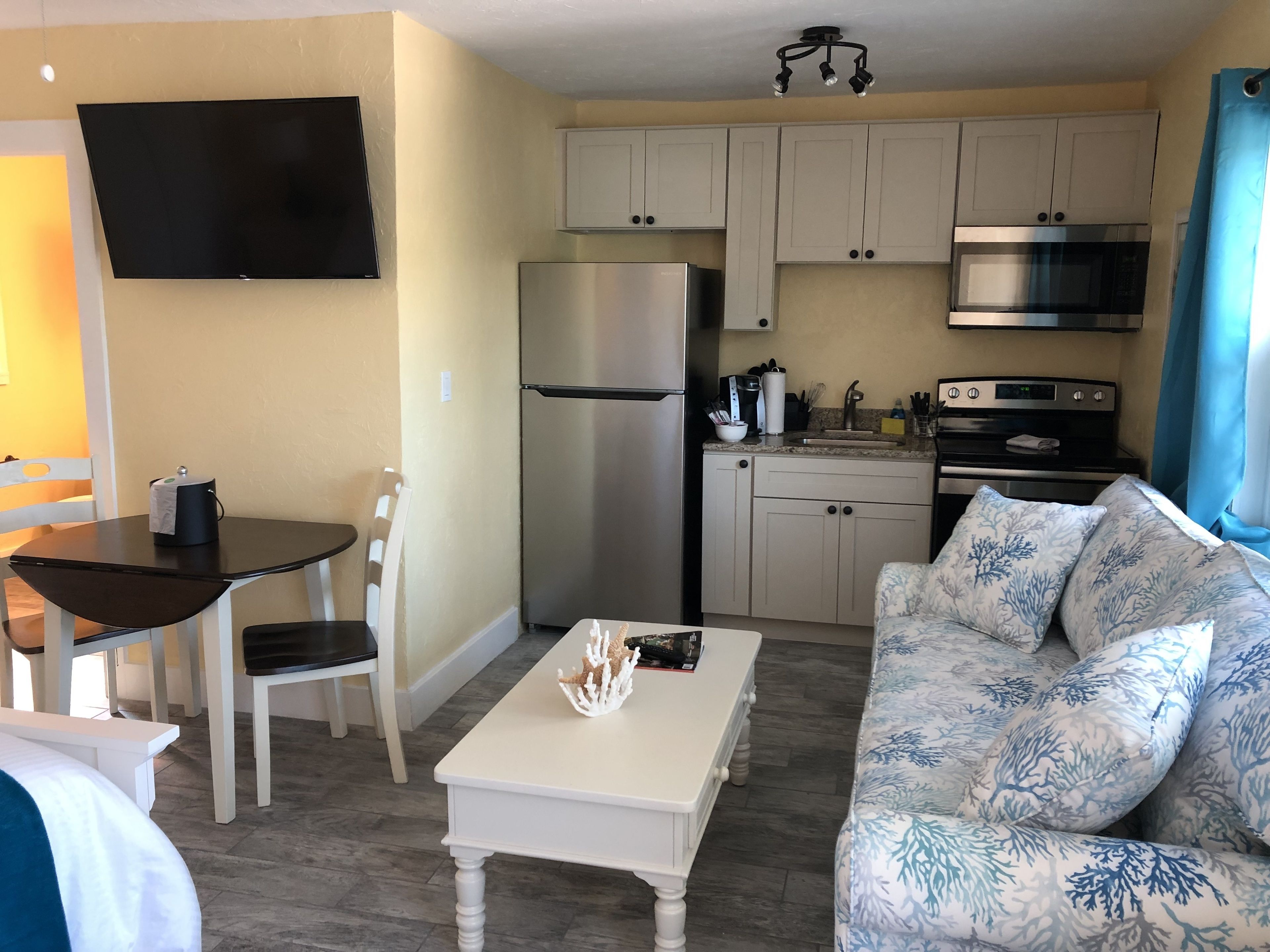 A compact living space with a dining table, a sofa, a kitchen area including a refrigerator, microwave, and stove, and a flat-screen TV mounted on the wall.
