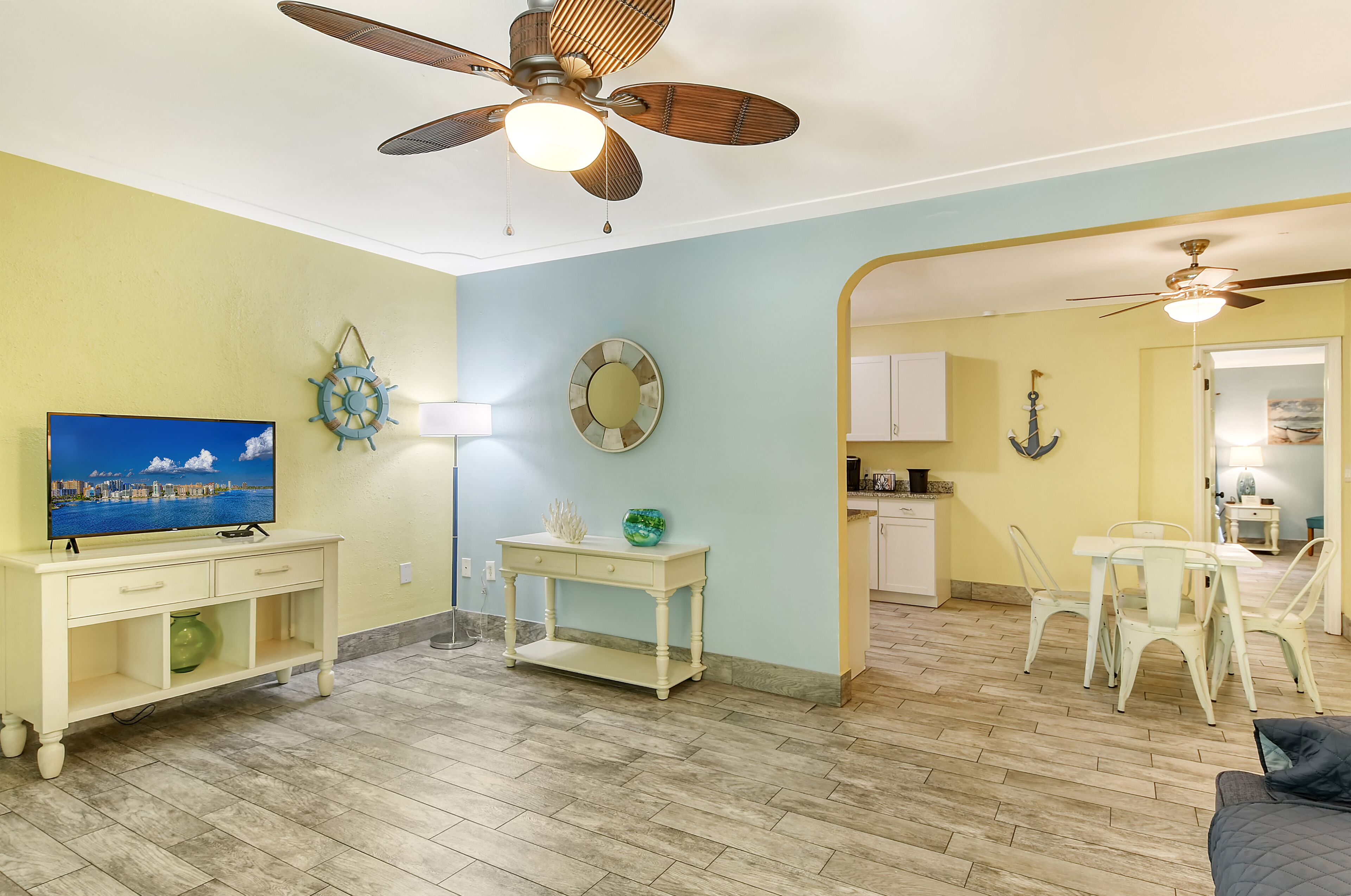 A living room with a television, a ceiling fan, a dining area with chairs, and a kitchen in the background.