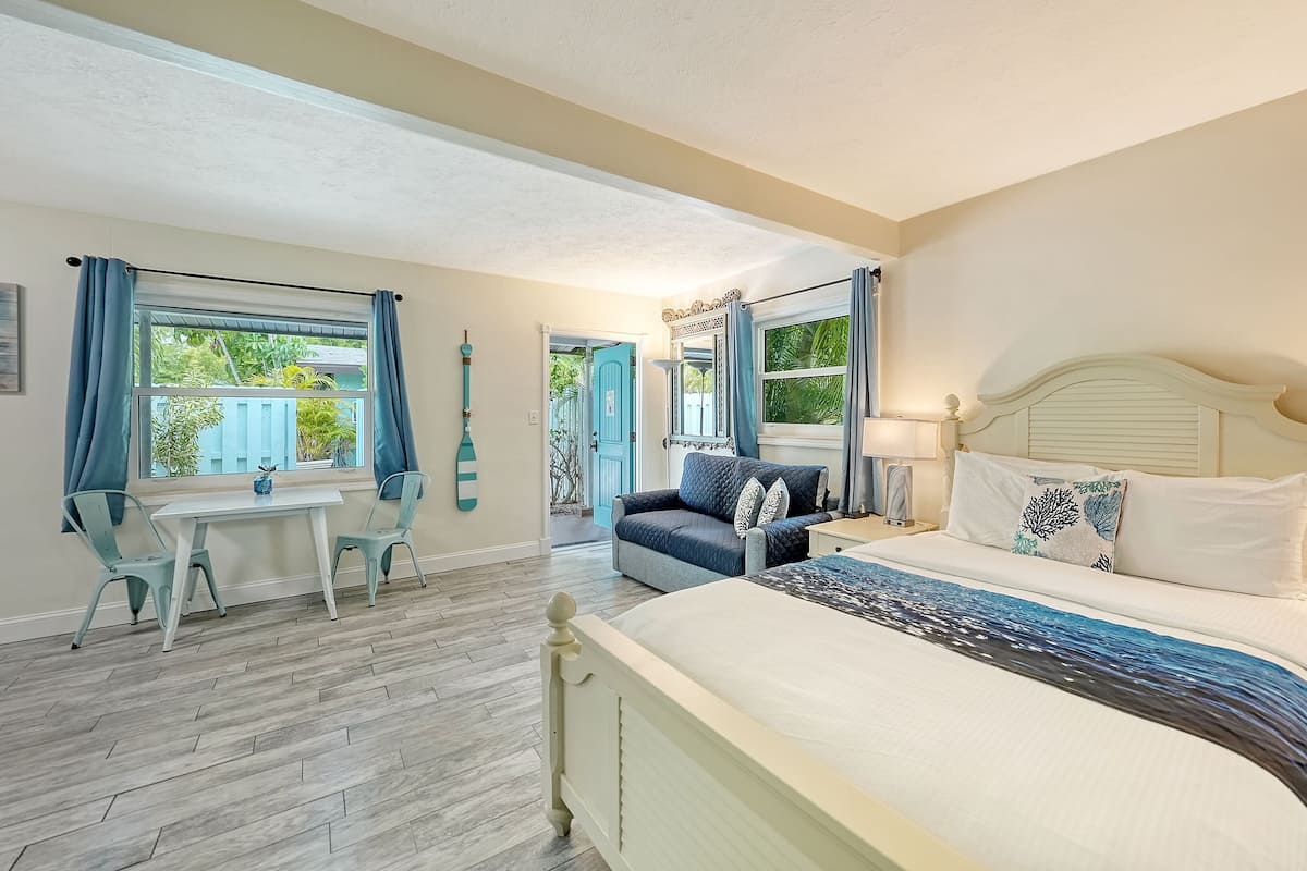 Un dormitorio con una cama grande, un escritorio y una silla. Hay ventanas con cortinas, una puerta y vistas al verde exterior.