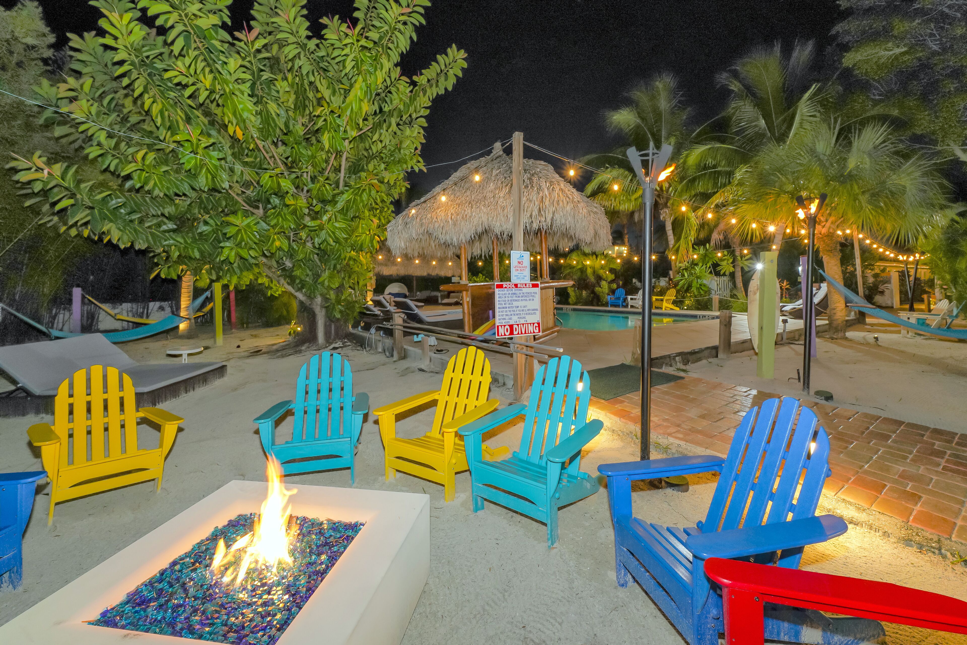 A poolside area with colorful Adirondack chairs, a fire feature, and a thatched-roof structure.