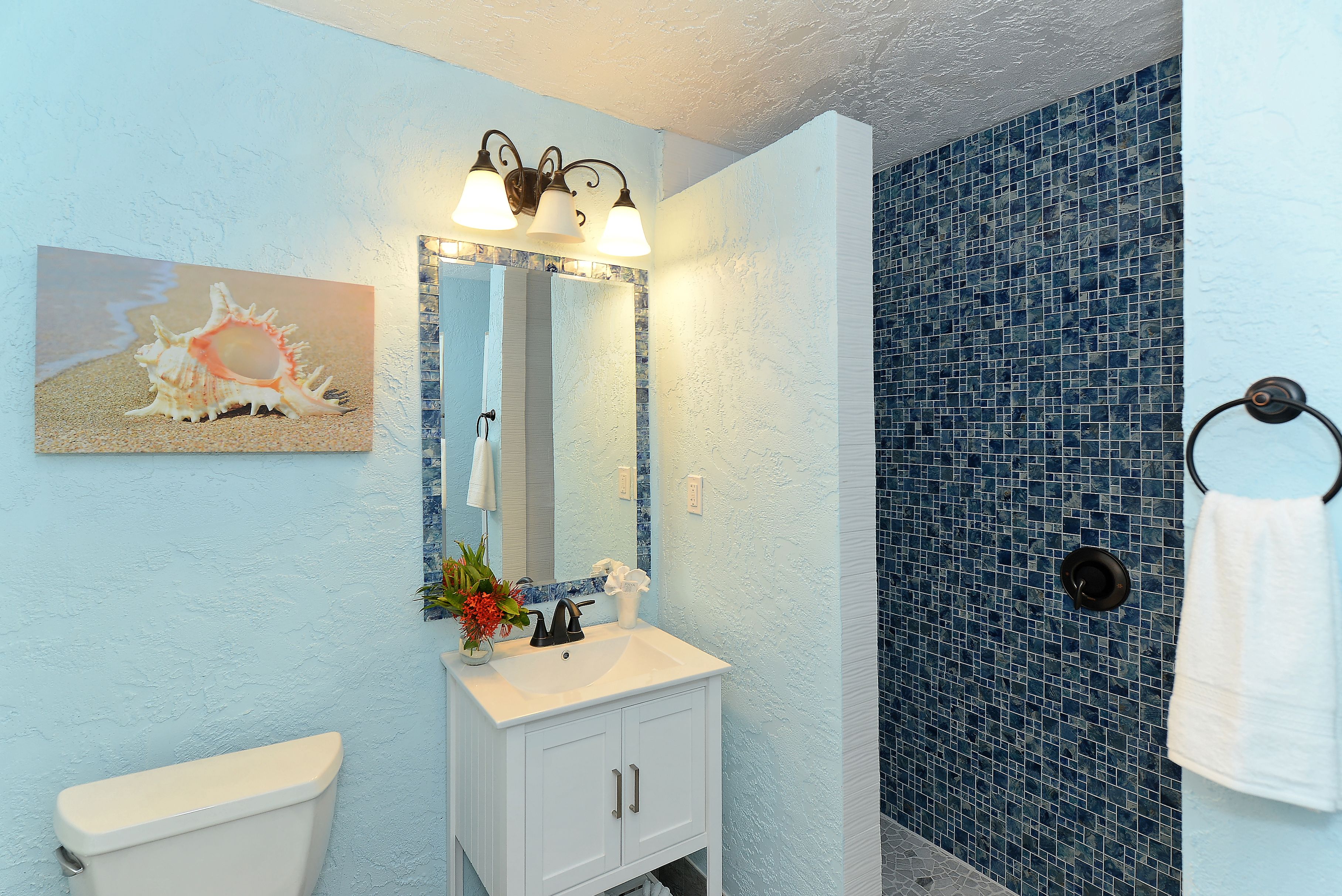 A bathroom with a white sink, a mirror with lights, a toilet, and a blue tiled shower wall.
