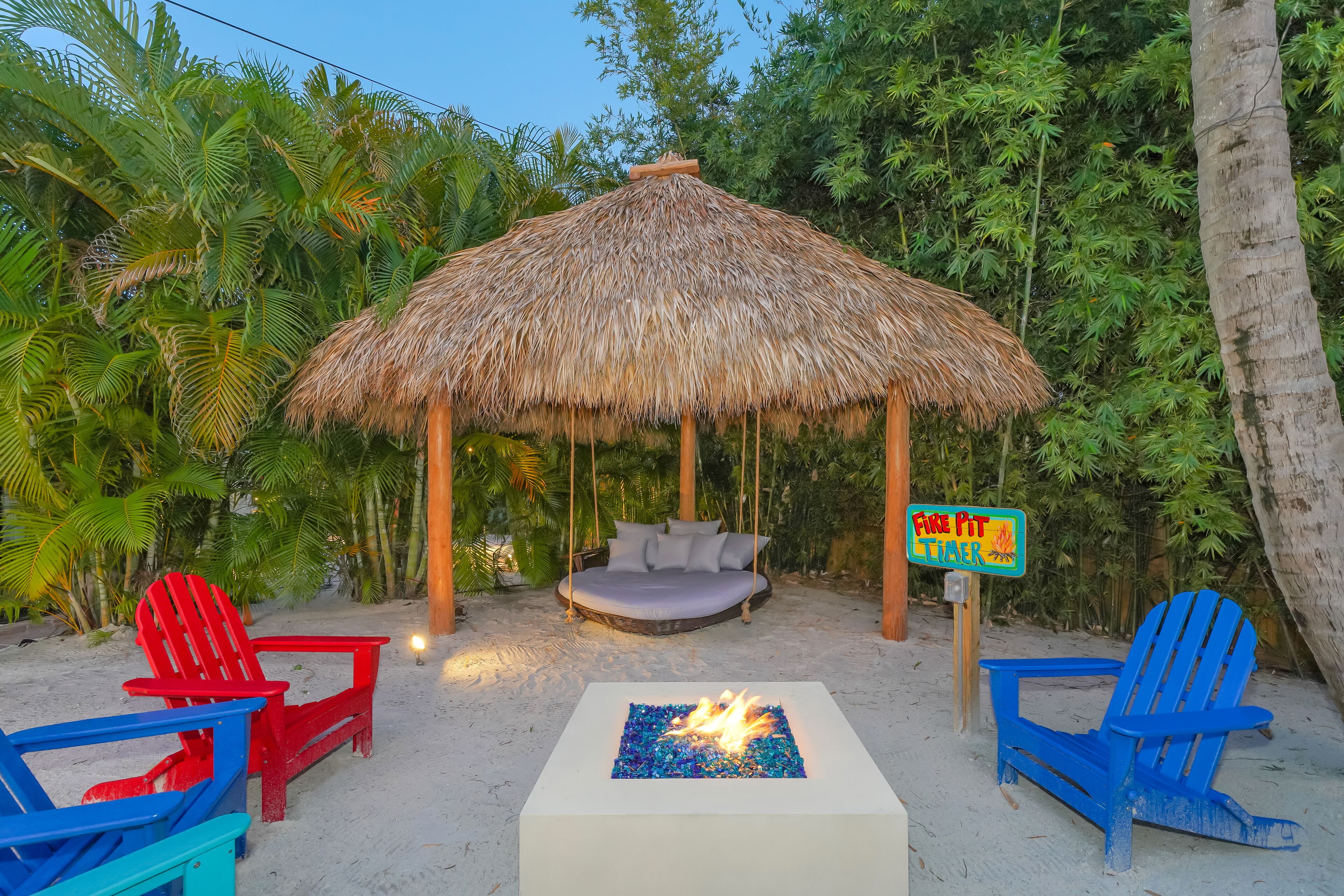 Eine Strandlandschaft mit einer strohgedeckten Struktur, einer Hängematte und einem Feuerstellenbereich mit Adirondack-Stühlen.