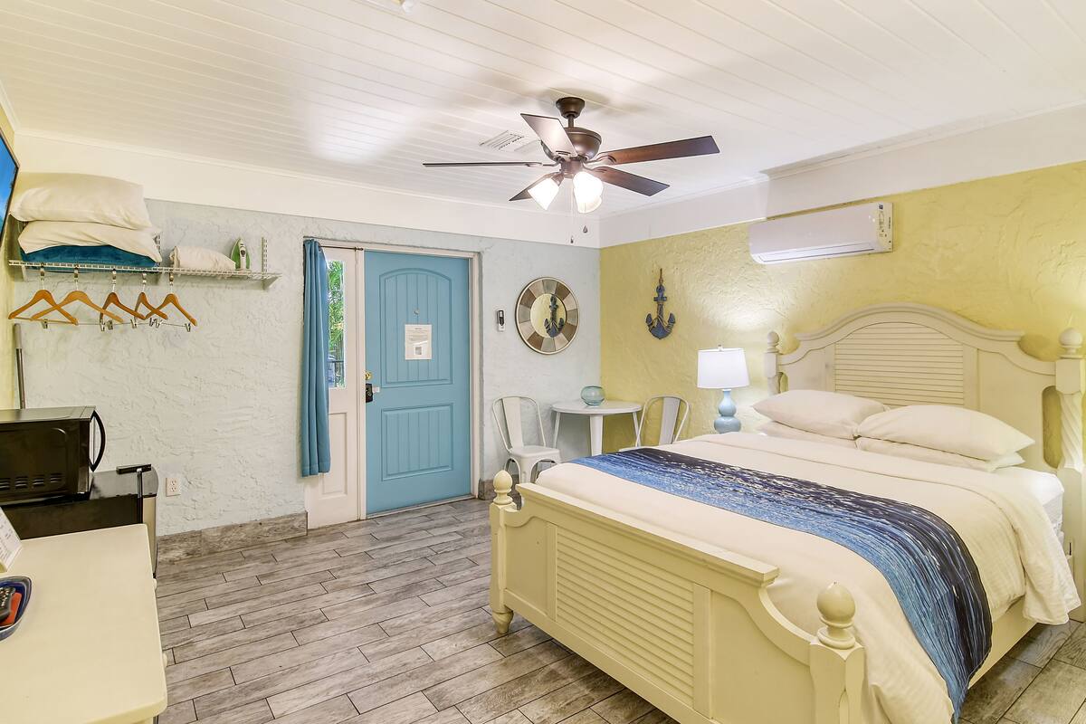 Un dormitorio con una cama, ventilador de techo, una pequeña mesa con sillas, microondas y una puerta azul.