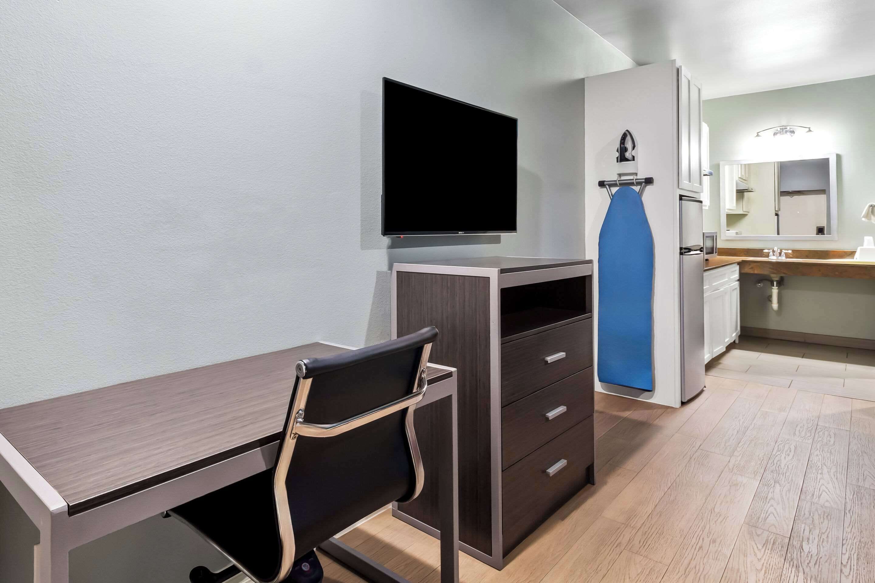 Een moderne hotelkamer met een flatscreen televisie aan de muur, een bureau met stoel, een kledingkast en een badkamer met wastafel en spiegel.