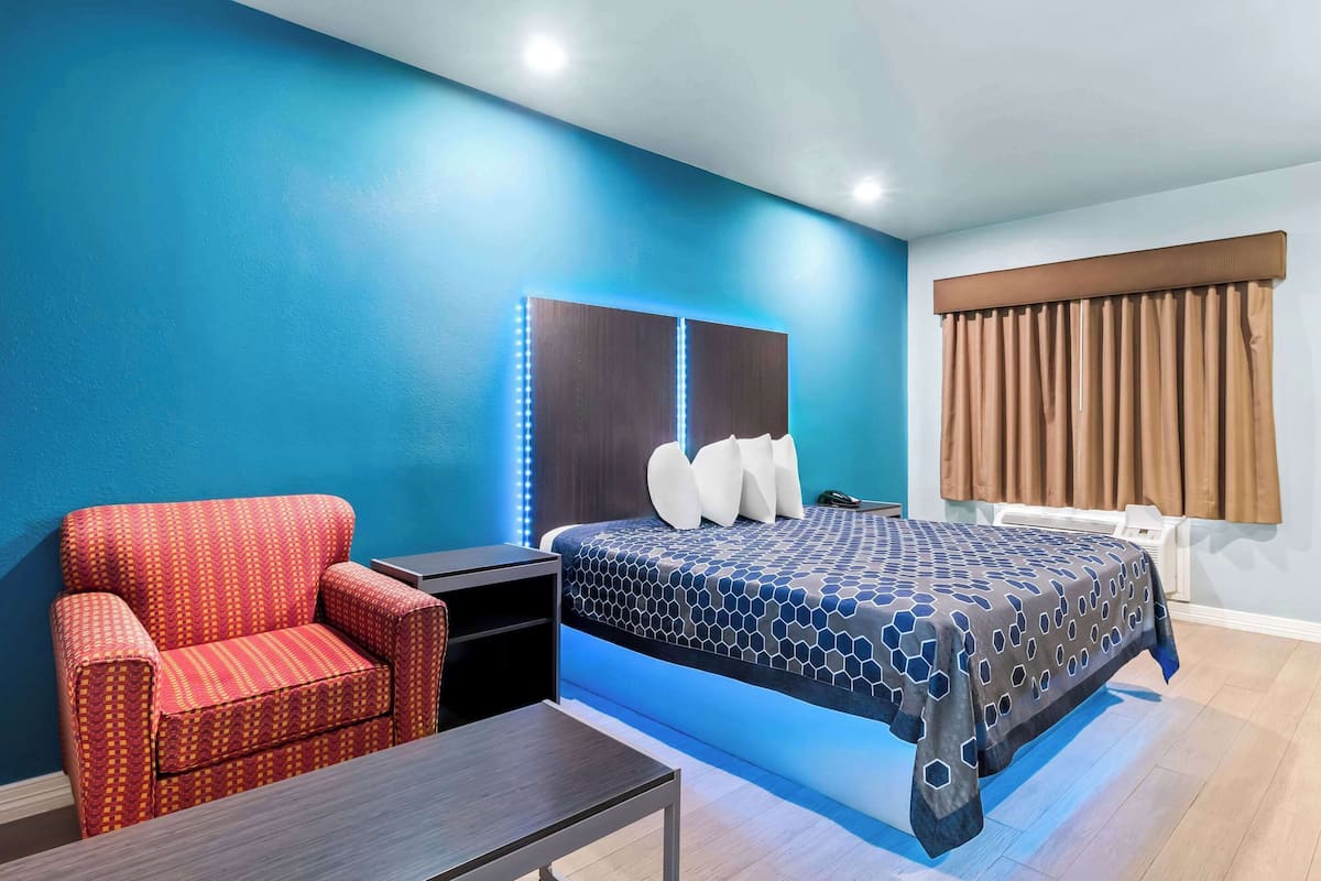 A hotel room with a blue wall, a bed with a patterned bedspread, a red armchair, a small table, and a nightstand.