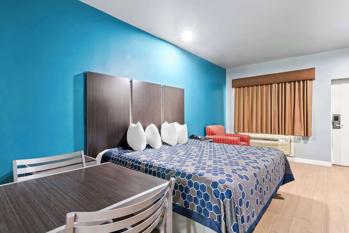 A hotel room with a blue wall, a wooden headboard, a bed with a patterned bedspread, a wooden table with chairs, and a window with brown curtains.