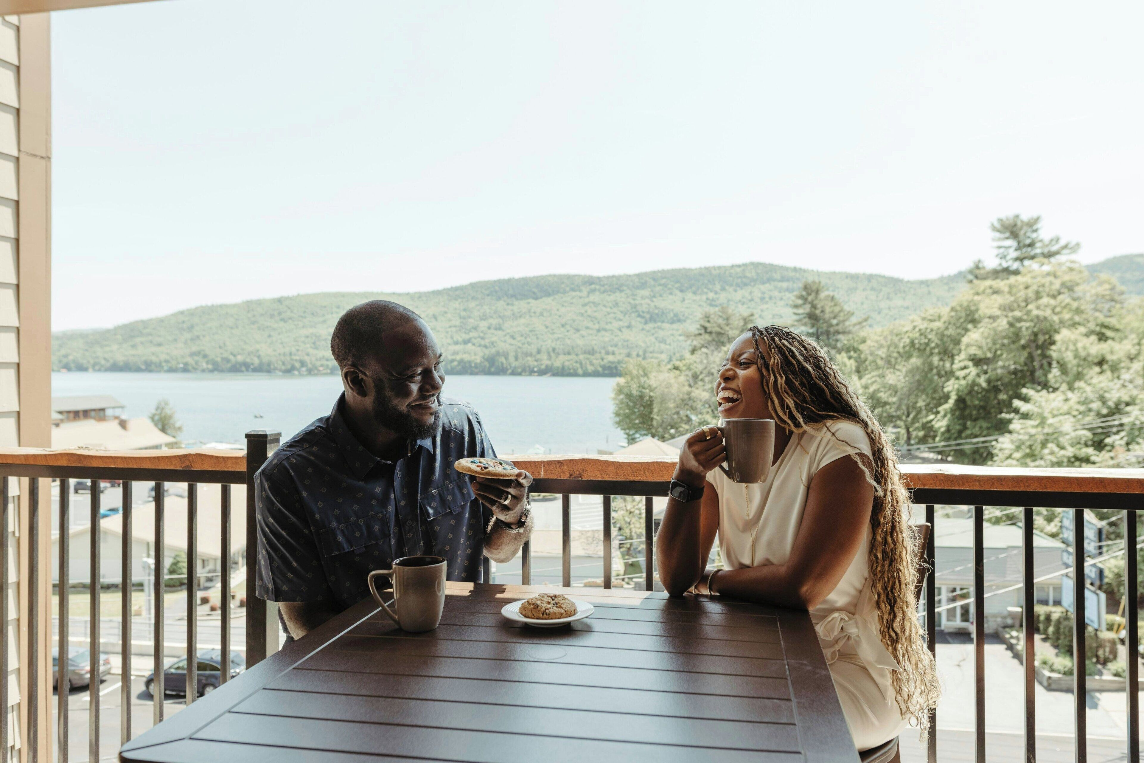 A couple sitting at a wooden table on a balcony overlooking a lake and hills.