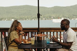 Dos personas cenando en una mesa al aire libre con pizza y bebidas, con vistas a un lago con un bote y colinas al fondo.