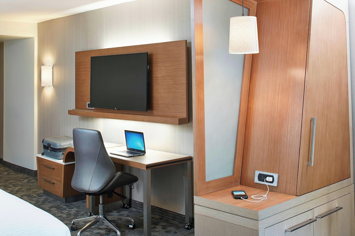 Habitación de hotel con escritorio de madera, televisor de pantalla plana, laptop, silla, armario y lámpara.