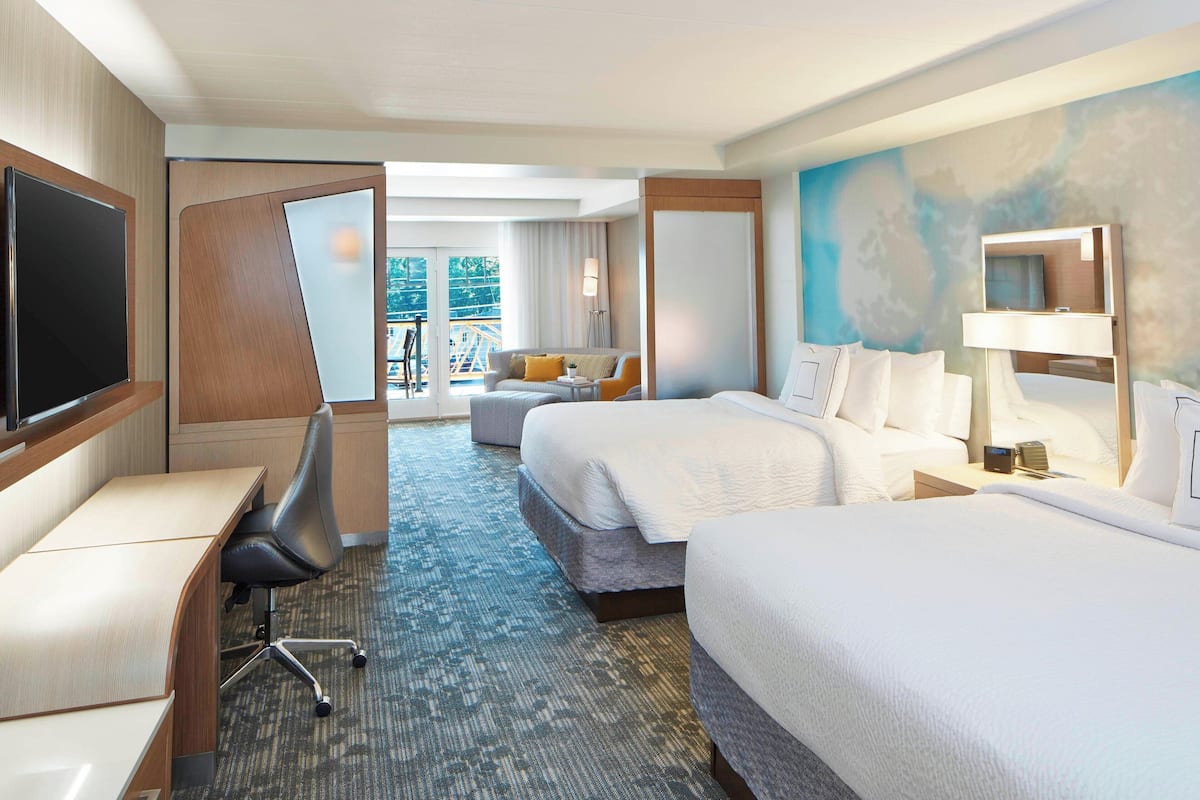 A hotel room with two beds, a desk, a chair, a TV, and a balcony with a view of trees.