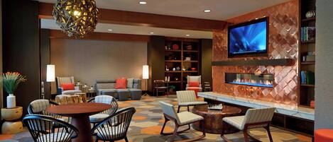 A modern lounge area with a round table, chairs, a fireplace, and a television mounted on a textured wall.