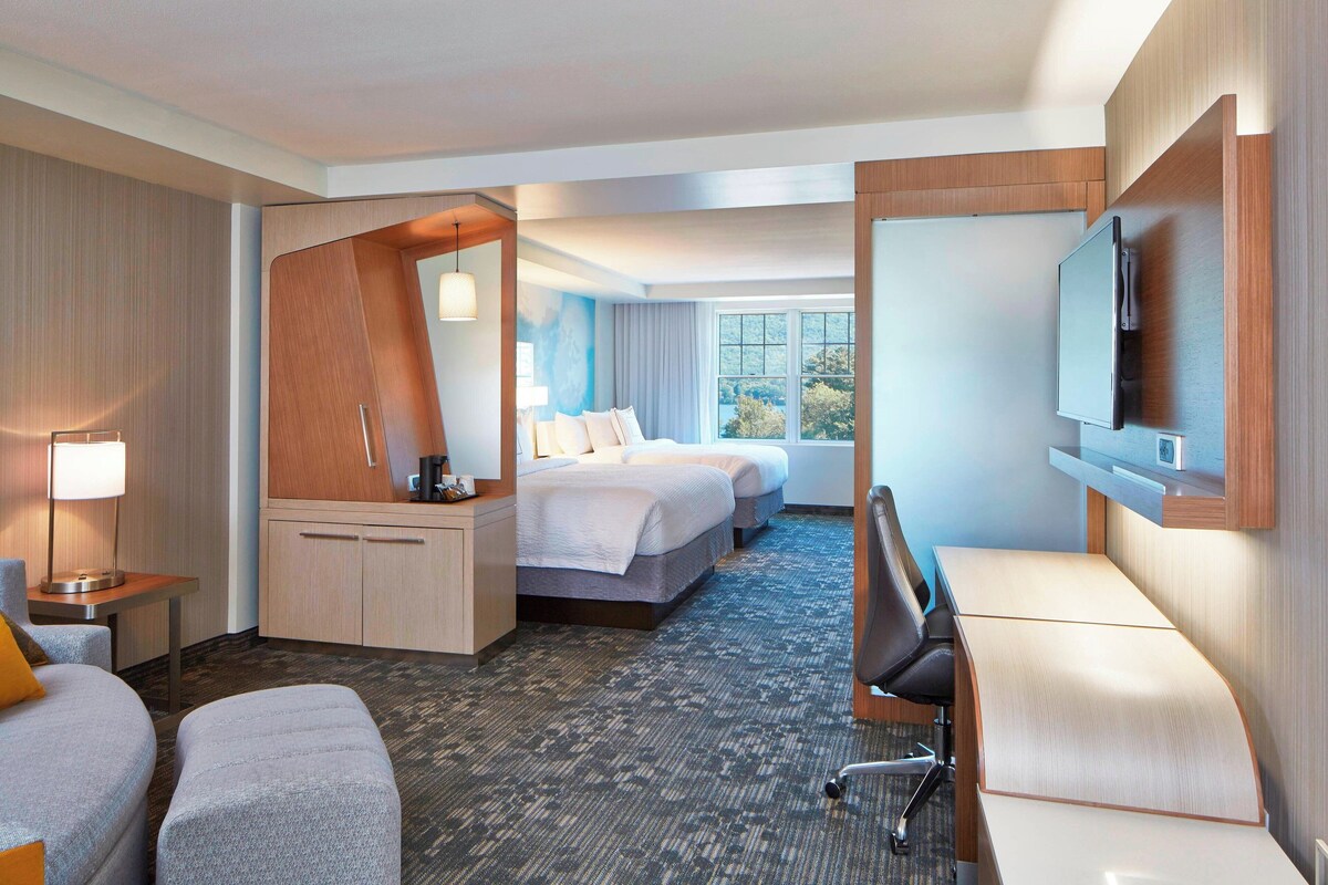Habitación de hotel con cama, escritorio, silla, televisor y ventana con vista a árboles.