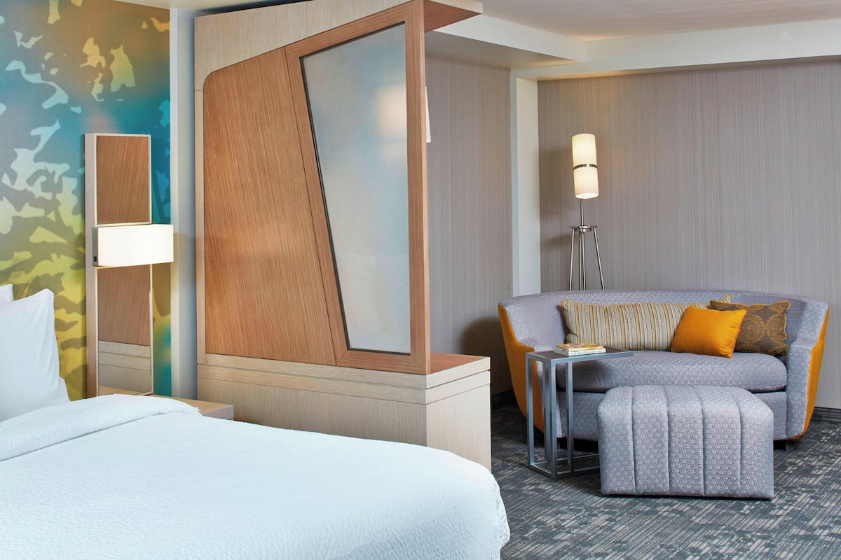 A hotel room with a bed, a sofa, an ottoman, a lamp, and a mirror.