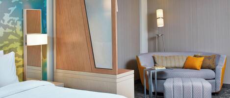 A hotel room with a bed, a sofa, an ottoman, a lamp, and a mirror.